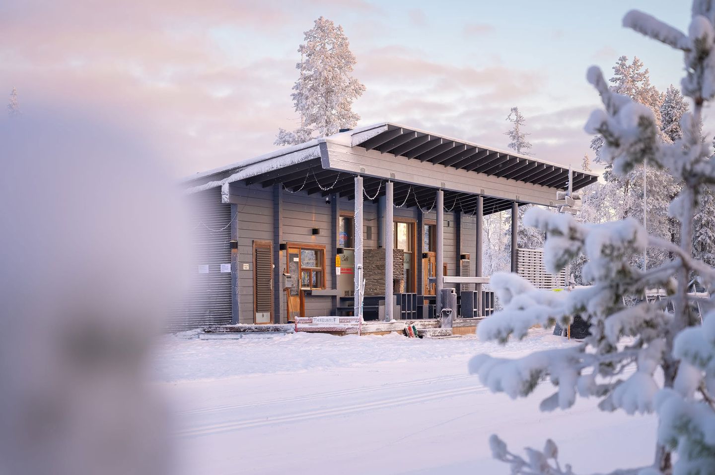 Uuttusuon kahvilan harmaa puurakennus lumen keskellä talviaamun vaaleanpunaisessa valossa, edessä latu.